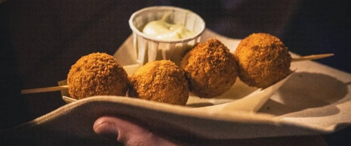 Quattro polpette di pesce impanate su uno spiedino di legno, con salsa bianca in una coppetta di carta, tenute in mano