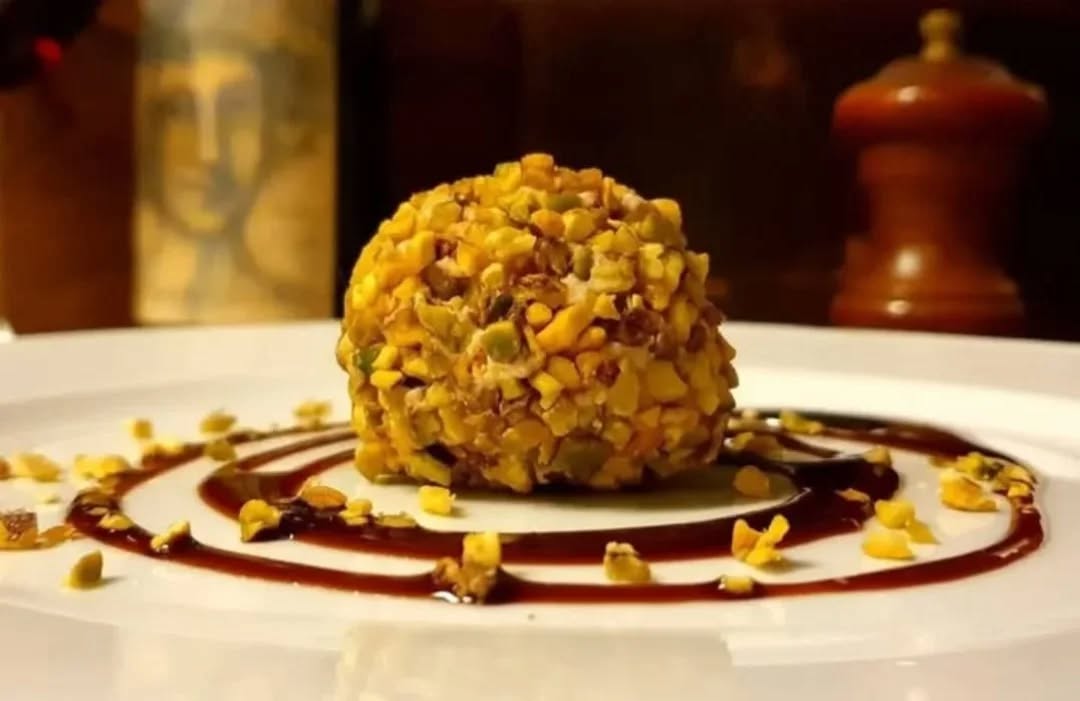 Polpetta dolce ricoperta di granella di pistacchio su spirale di salsa scura, piatto bianco de La Pecora Nera polpette & Co
